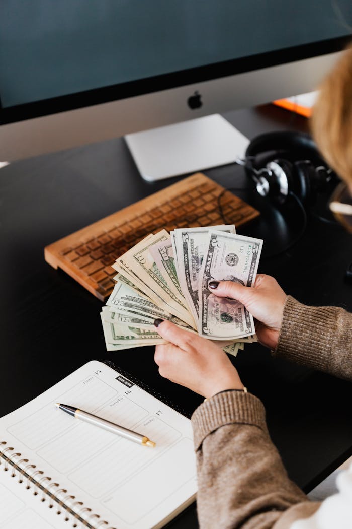 Close-up of hands counting dollar bills at a desk with computer and planner.